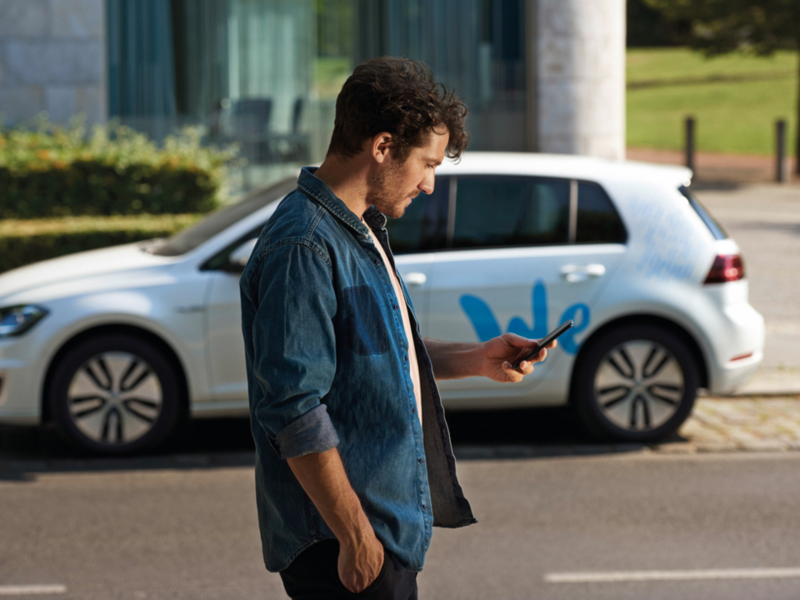 Hombre joven consultando un móvil delante de un Golf blanco aparcado con el logo de Volkswagen We