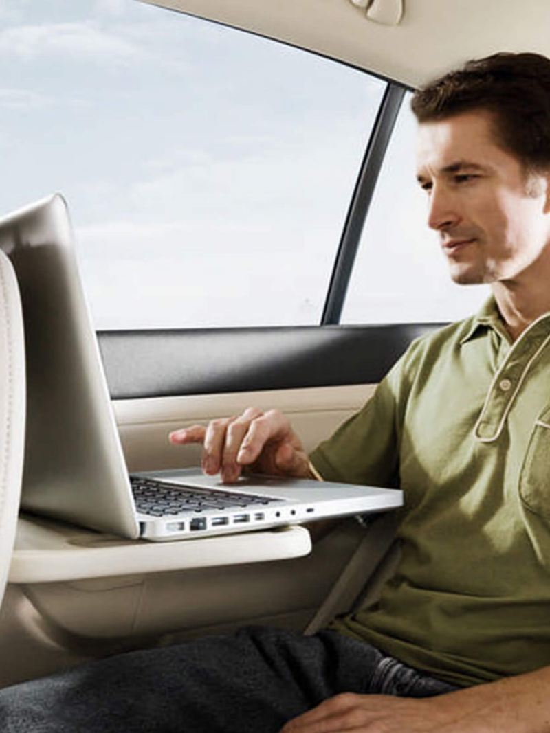 A man using his laptop on a foldout table in the back passenger seat of a Volkswagen Golf.