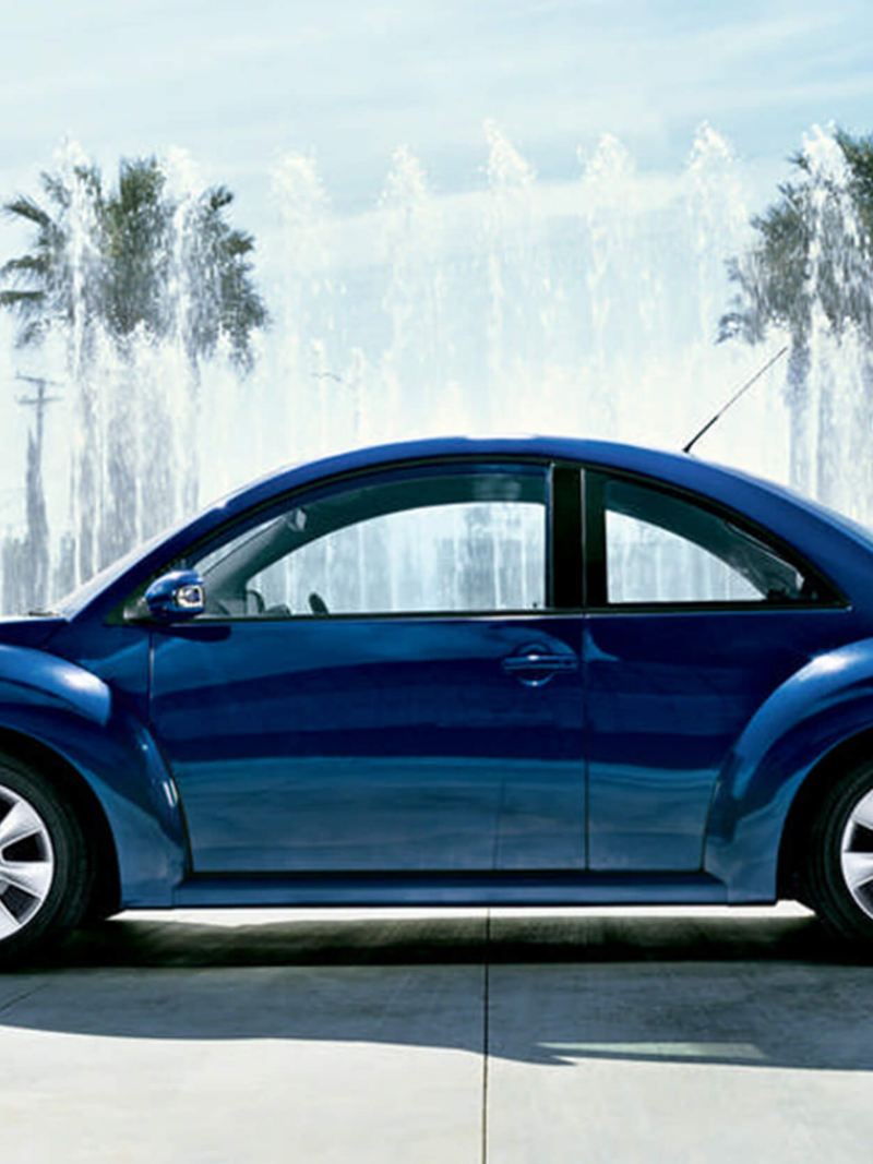 Profile shot of a blue Volkswagen Beetle, fountains and tree's in the background.