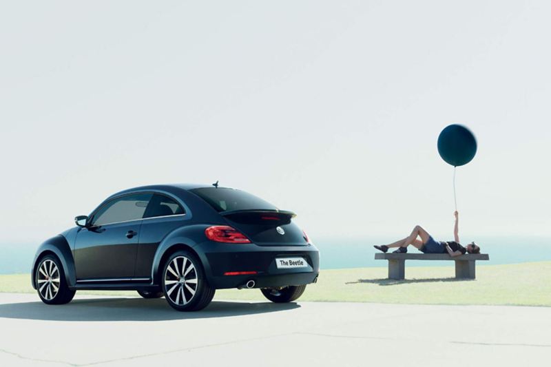 Rear shot of a black Volkswagen Beetle, a lady lay on a bench holding a balloon in the background.