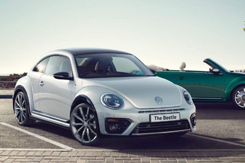 3/4 front view of a white Volkswagen Beetle, with a green Volkswagen Beetle Cabriolet in the background.