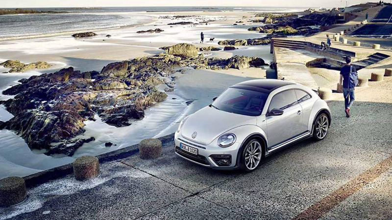 A white Volkswagen Beetle, on a rocky coastal road.