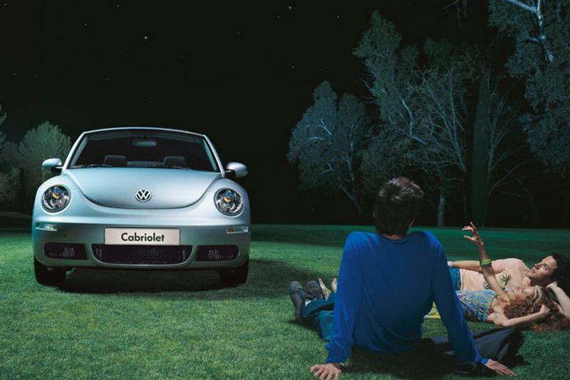 A blue Volkswagen Beetle Cabriolet, in a field with a couple under a tree.