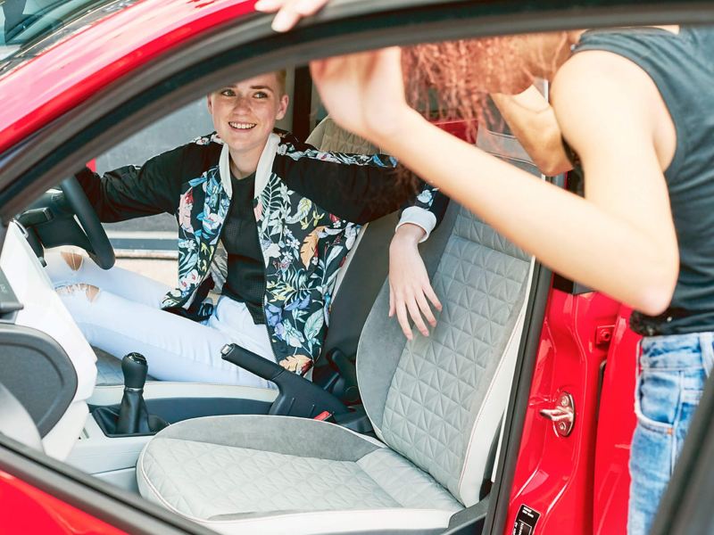 Two ladies one in the drivers seat, the other at the open passenger door of a red Volkswagen up!