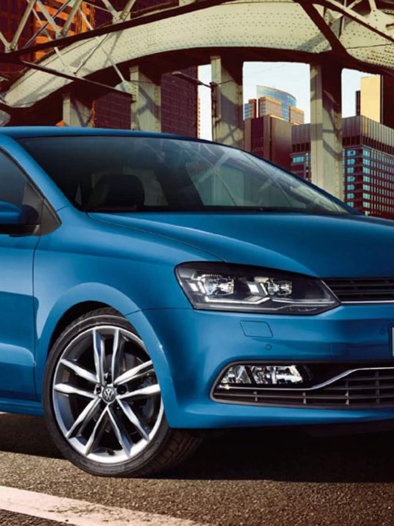 A blue Volkswagen Polo, under a steel structure, in a city setting,