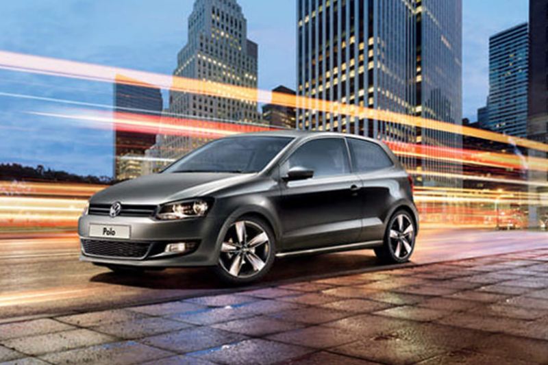 A silver Volkswagen Polo, driving through a city, skyscrapers in the background.