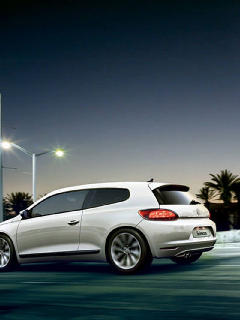 Rear profile shot of a white Volkswagen Scirocco, at dusk with palm trees in the background.
