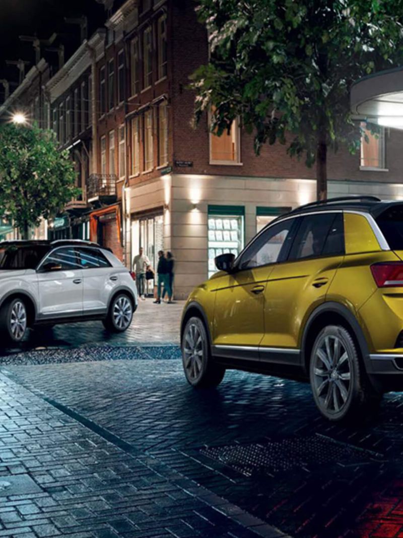 A white and a yellow Volkswagen T-Roc, on a city street at night.