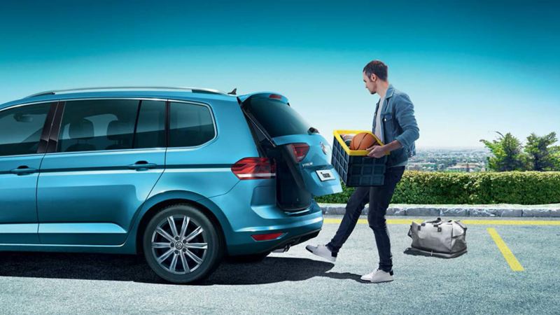 A man with a box  using the hands-free sensors to open the boot of a blue Volkswagen Touran.