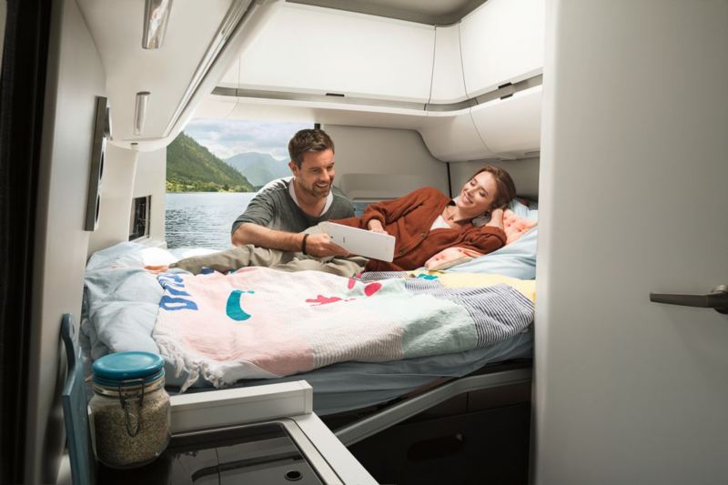 Mężczyzna i kobieta oglądają film leżąc na łóżku w Grand Californii, w tle otwarte drzwi z widokiem na jezioro i góry
