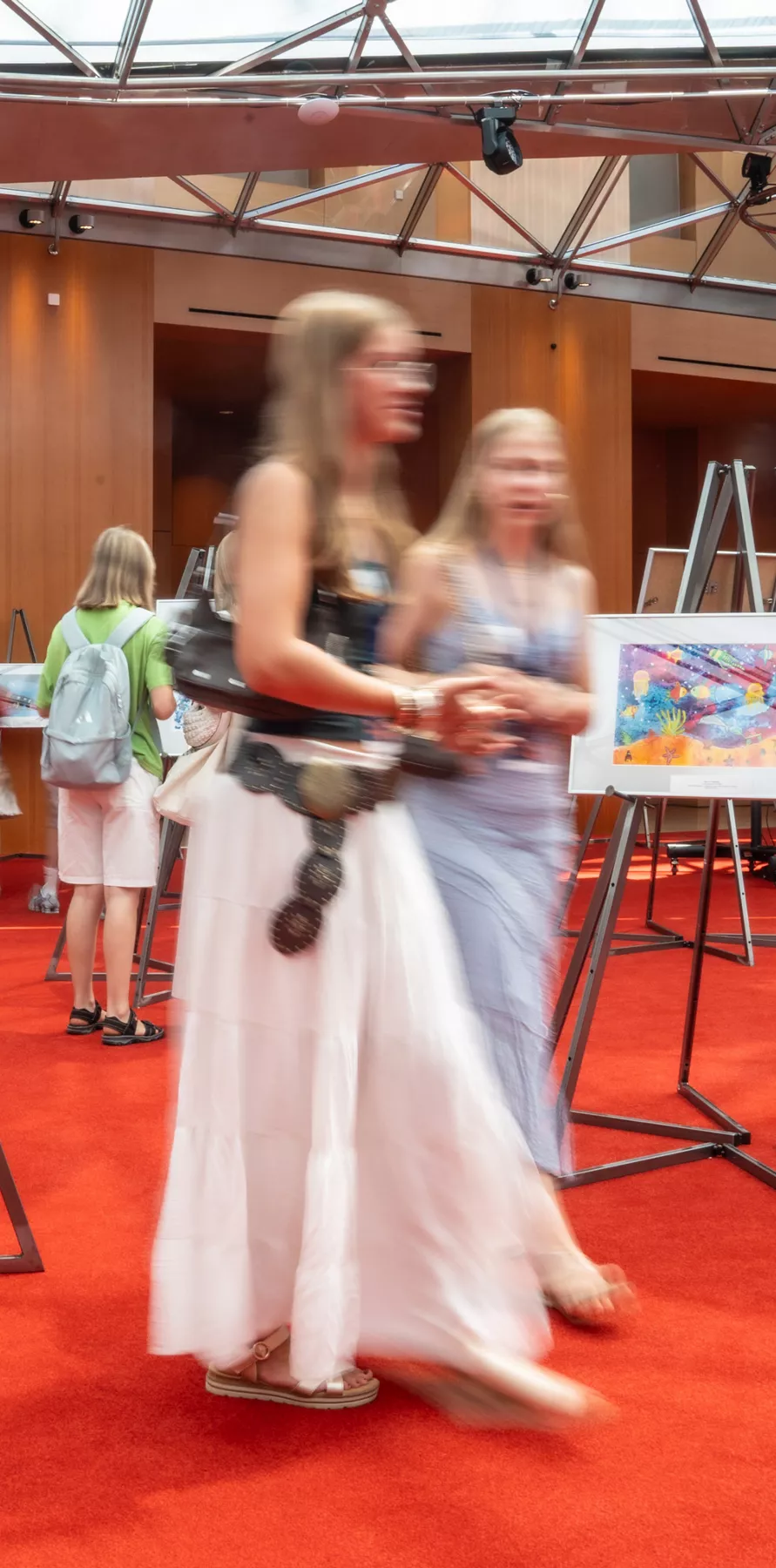 Besuchende betrachten auf Staffeleien präsentierte Bilder in einer Ausstellungshalle mit rotem Teppich.
