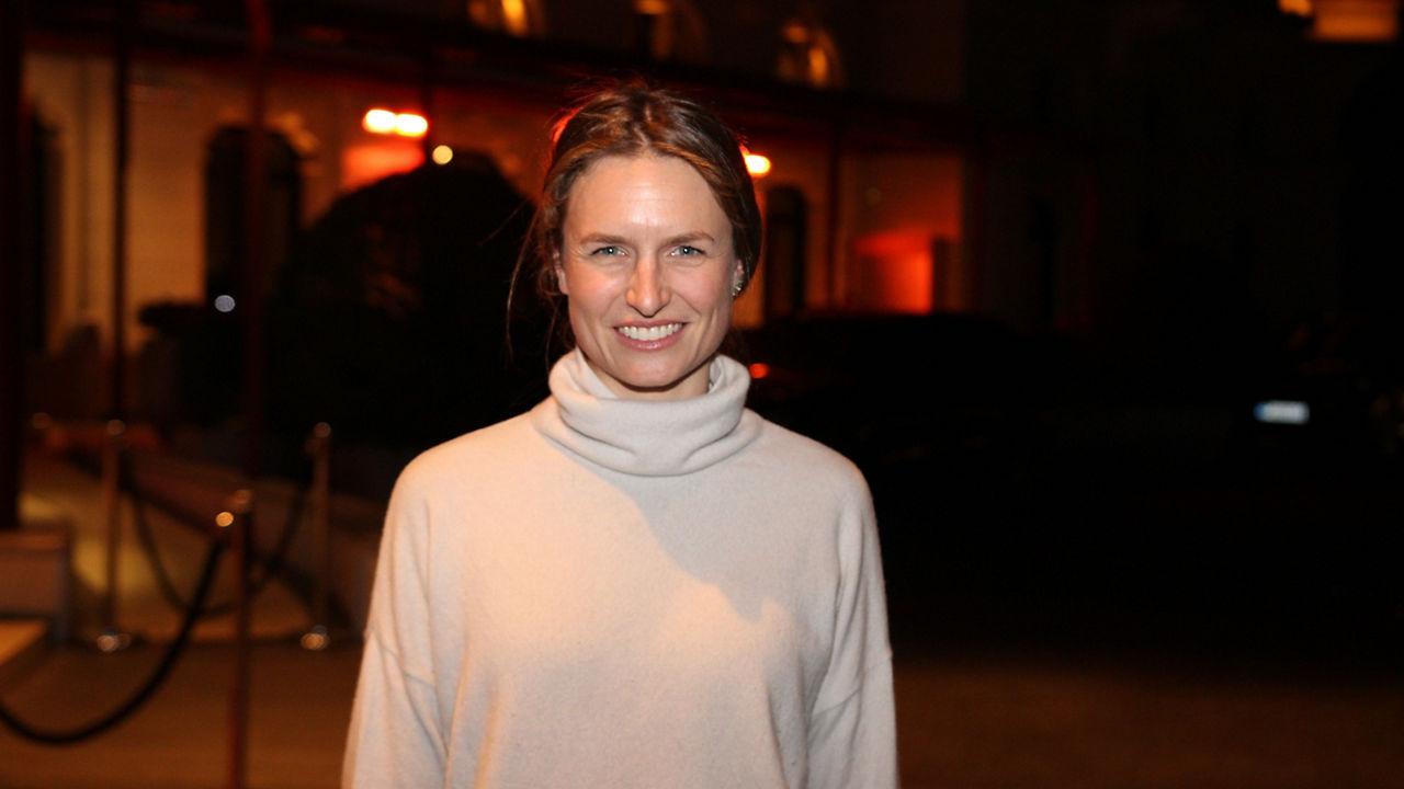 Eine junge Frau in Rollkragenpullover blickt lachend in die Kamera. Sie steht in einem Raum, der nur spärlich  mit rotem Licht erleuchtet wird.