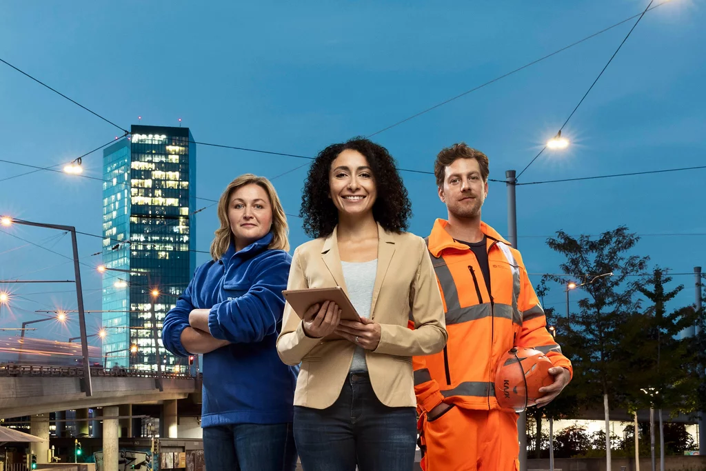 Drei Mitarbeitenden vom erz in Arbeitskleidung in Dämmerung vor Prime Tower stehend.