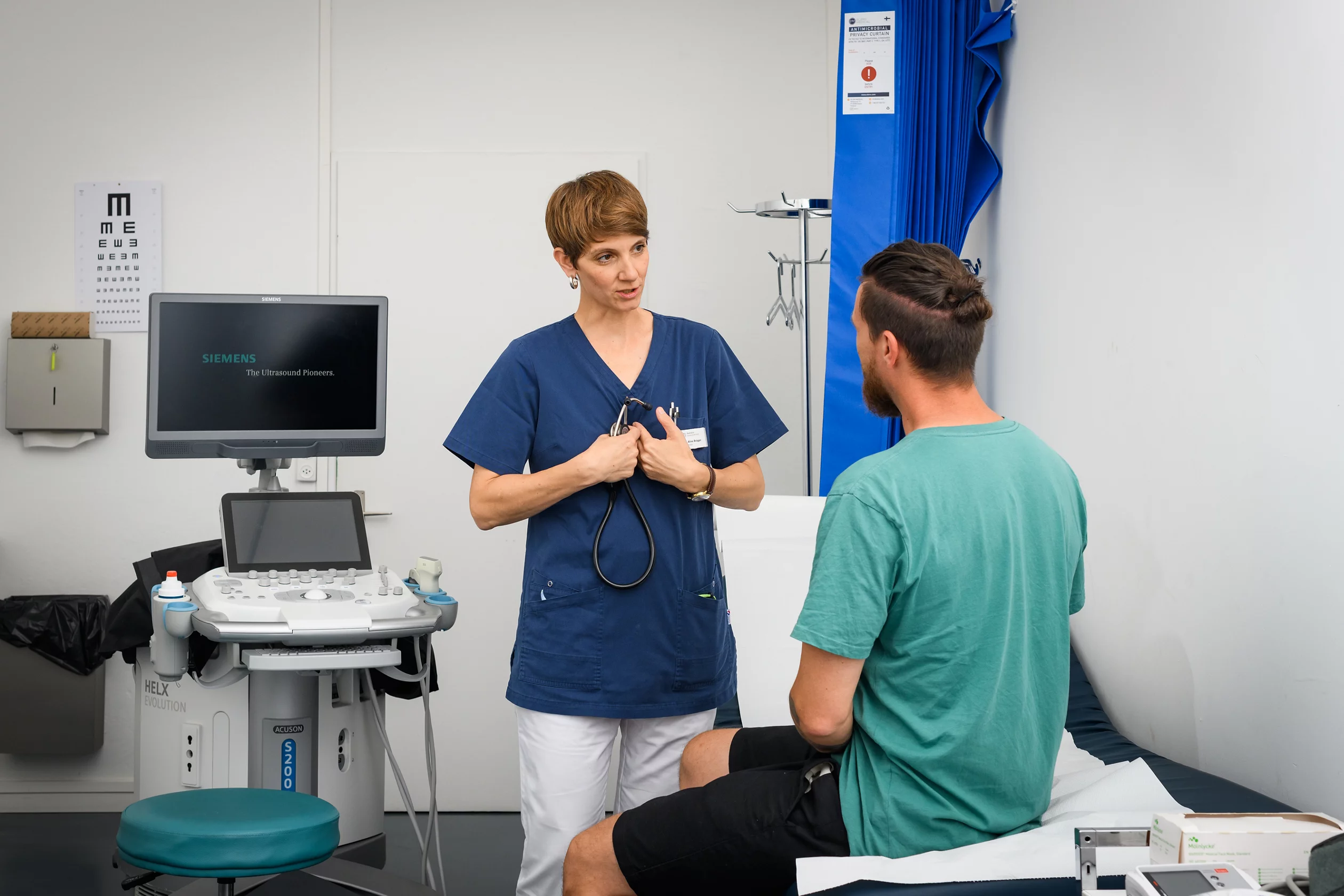 Gespräch in Untersuchungsraum zwischen Ärztin in blauer Arbeitskleidung und Patient in grünem T-Shirt auf Liege sitzend.