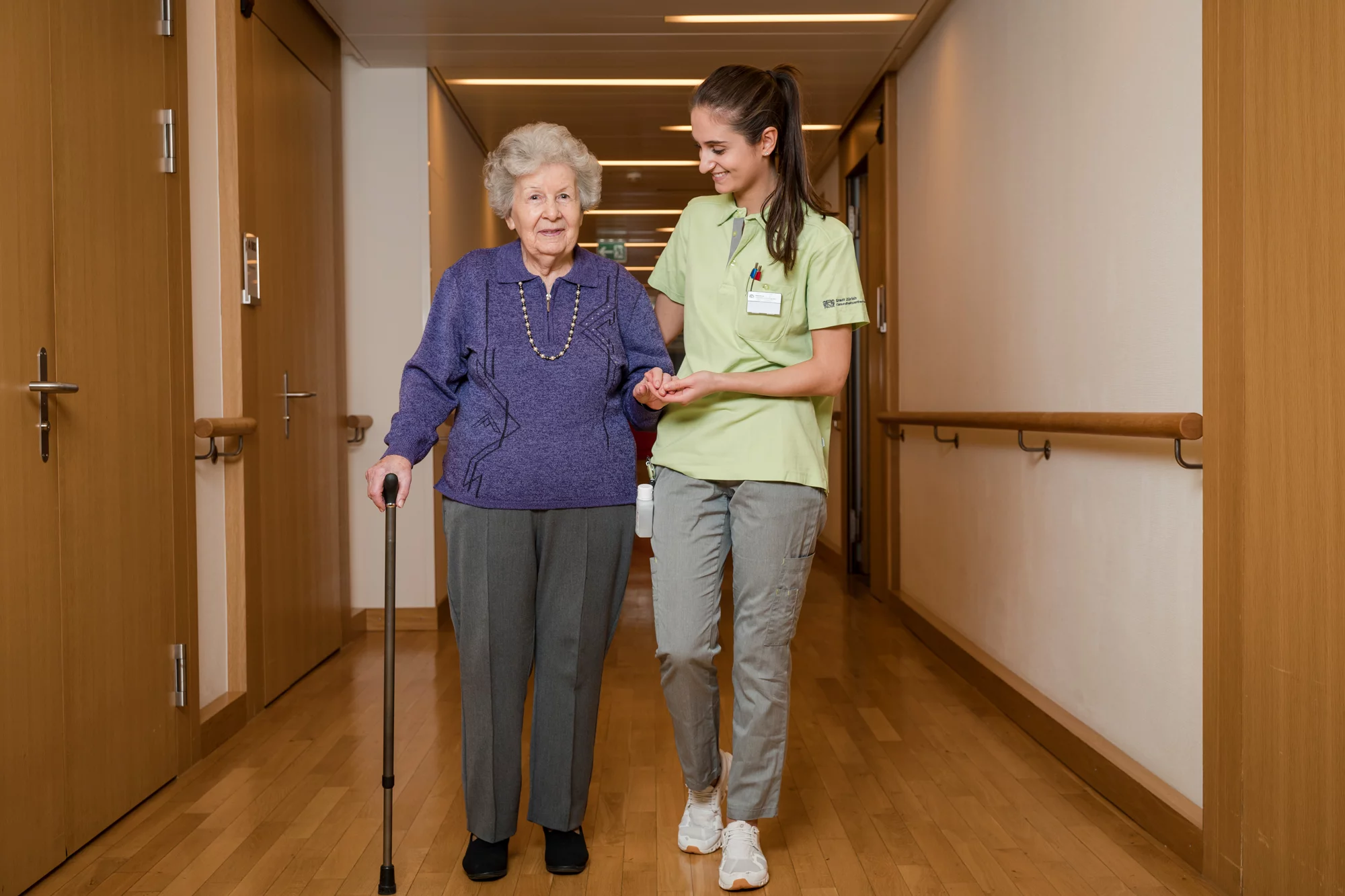Assistentin Gesundheit und Soziales in Ausbildung führt eine Bewohnerin den Flur entlang.