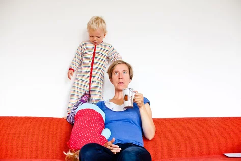 Eine Frau trinkt aus einer Tasse, während zwei Kinder um sie herumturnen.