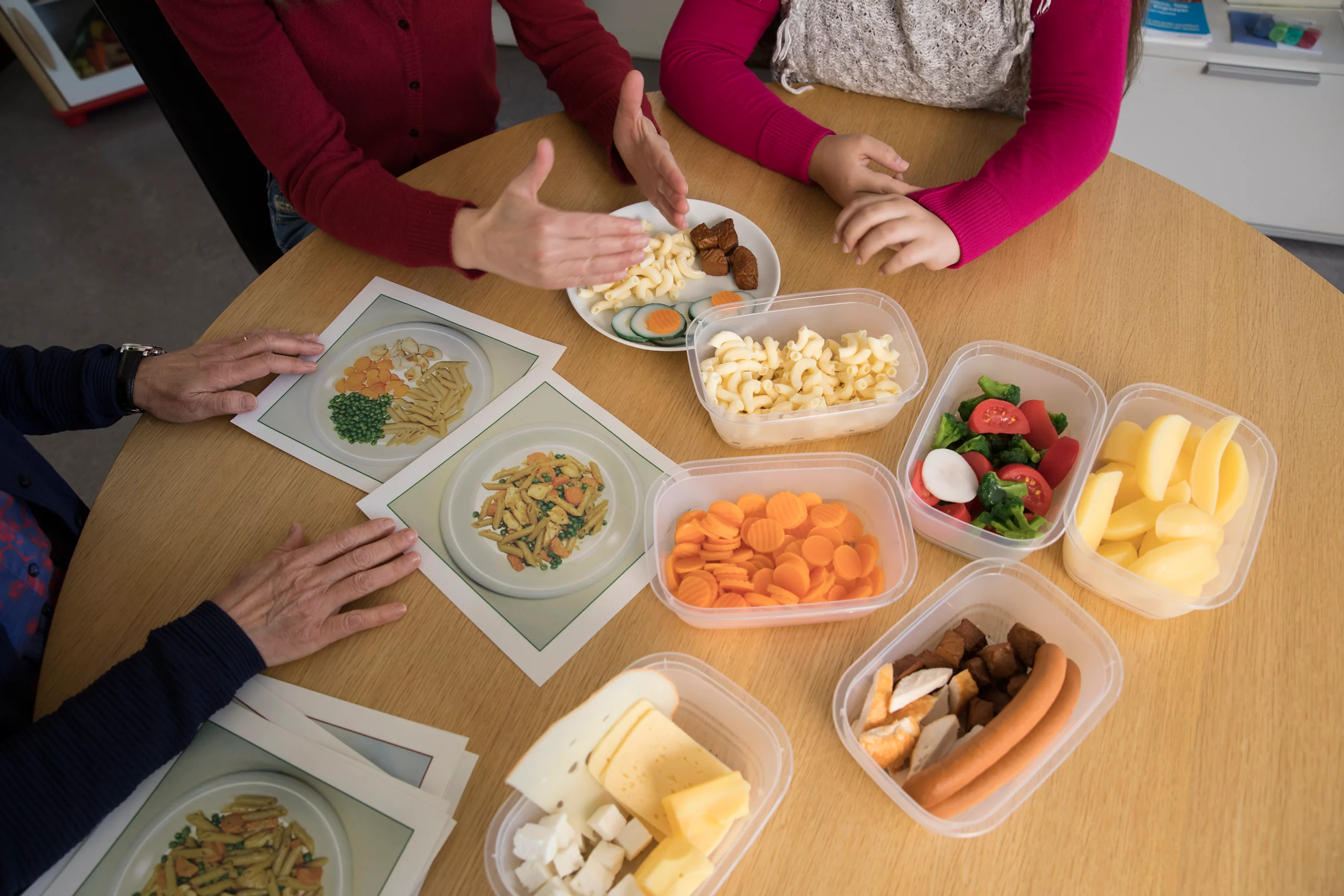 Ernährungsberaterin, Mutter und Tochter sitzen um einen Tisch und sprechen miteinander über Lebensmittel, welche auf dem Tisch sind.