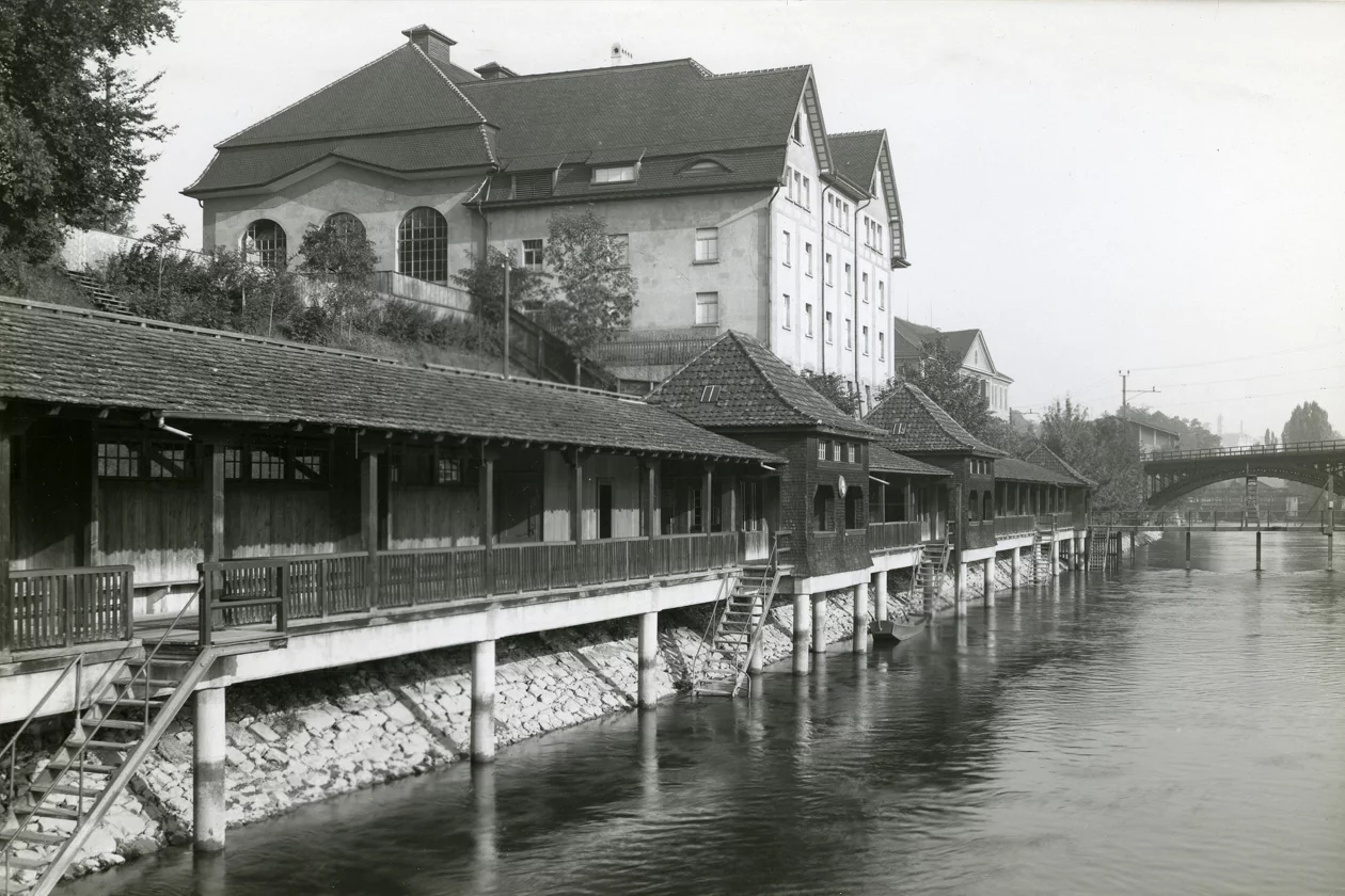 Historische Schwarzweissaufnahme des Flussbads Unterer Letten am Flussufer mit Treppen ins Wasser, grossem Gebäude am Hang und Brücke im Hintergrund.