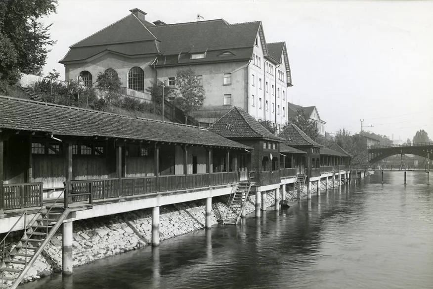 Historische Schwarzweissaufnahme des Flussbads Unterer Letten am Flussufer mit Treppen ins Wasser, grossem Gebäude am Hang und Brücke im Hintergrund.