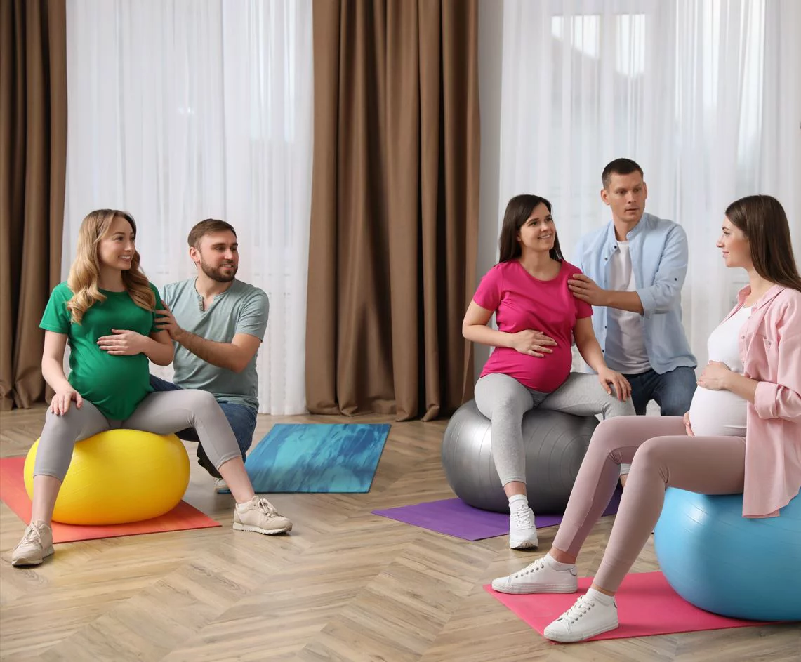 Geburtsvorbereitungskurs, drei Frauen auf einem Gymnastikball und ihre Männer helfen