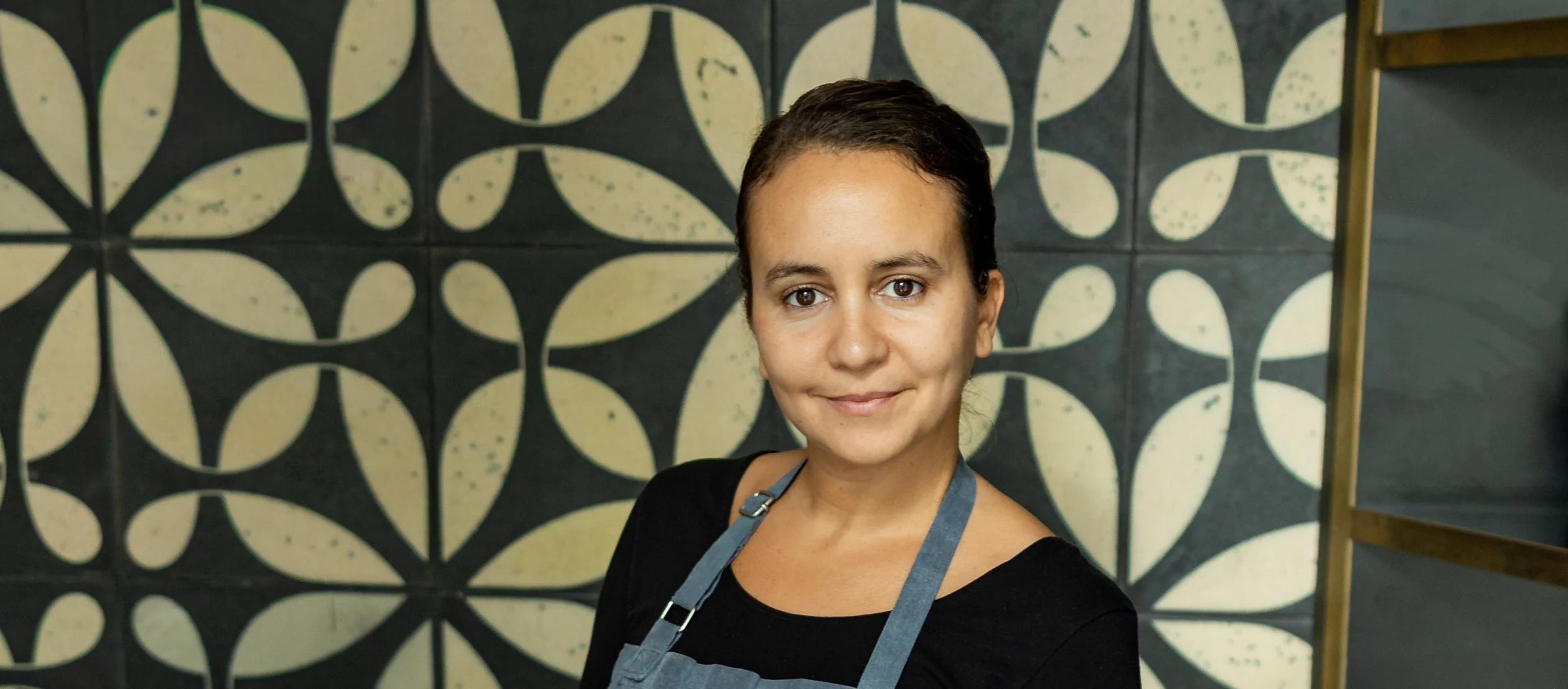 Eine junge Frau, die Haare zurückgebunden und mit einer Schürze, steht vor einer Tabette mit Ornamenten.