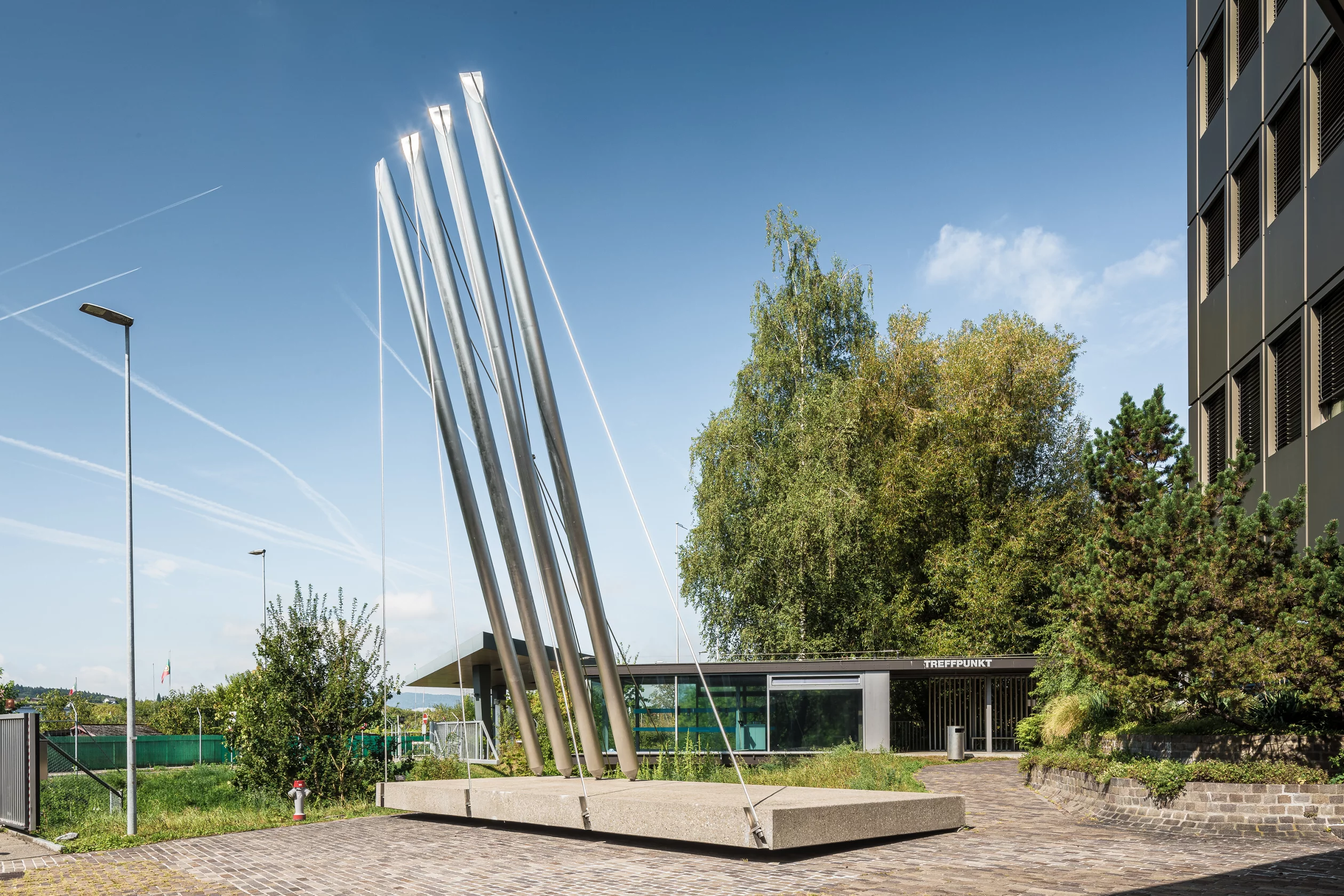 Vier in den Himmel ragende Masten aus Chrom, Nickel und Stahl; befestigt an Stahlseilen und Betonplatte