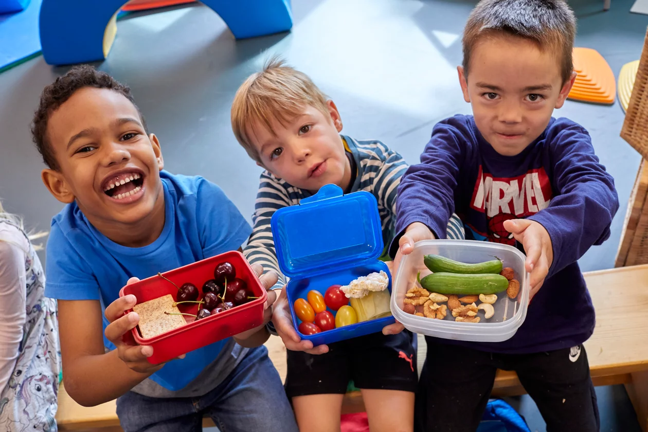 Drei Jungen zeigen im Kindergarten ihre gefüllten Znüniboxen