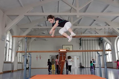 Ein Kind springt über den Turnbarren. Im Hintergrund hat es noch andere Kinder sich in der Turnhalle am bewegen.