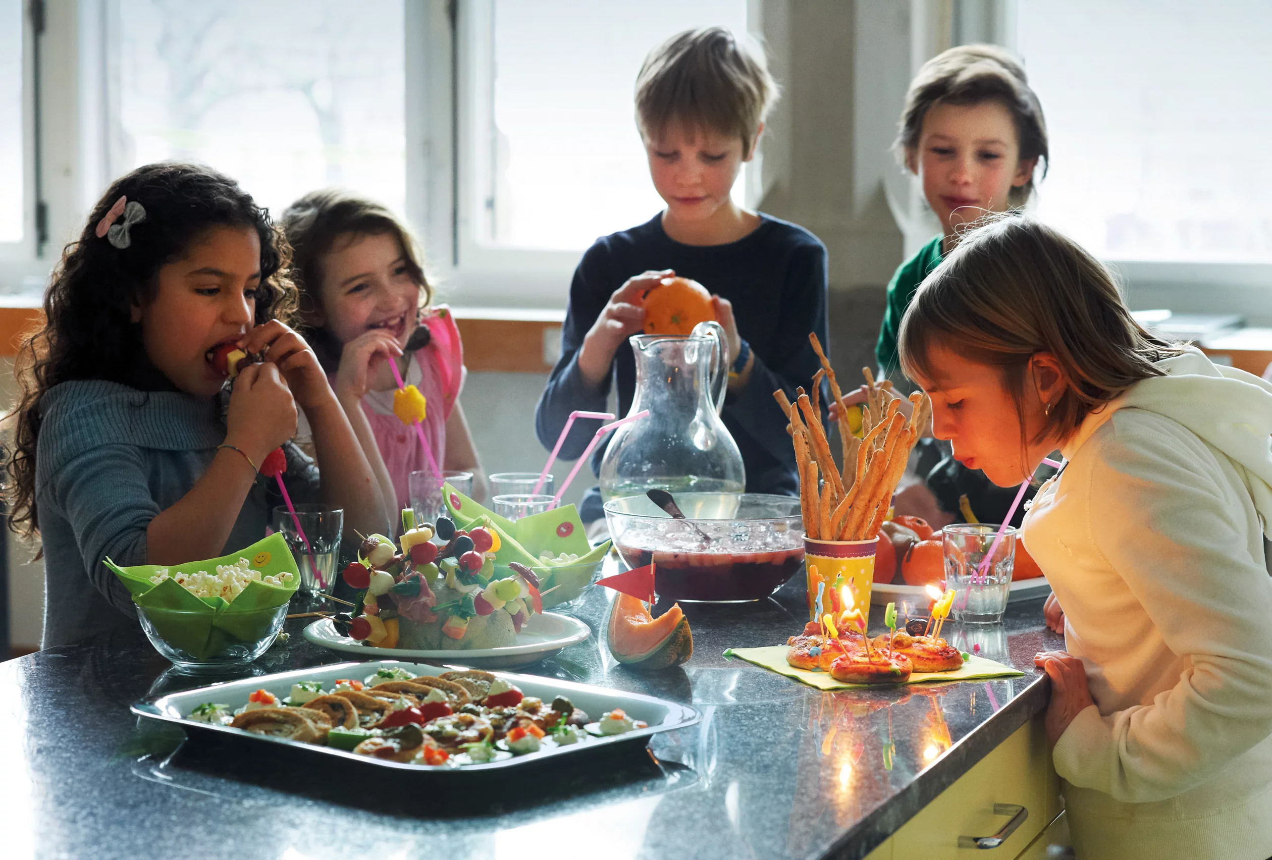 Fünf Kinder stehen um einen Tisch voller Apéro. Ein Kind bläst Kerzen aus.