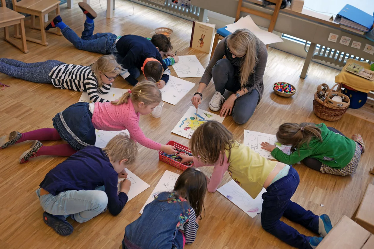Kindergarten-Kinder sitzen mit der Lehrperson am Boden und zeichnen auf Blätter
