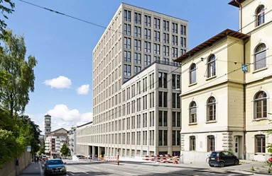 Der linke und der rechte Flügel des hellen, mehrstöckigen Gebäudes orientieren sich an der Höhe der benachbarten Gebäude. Der mittlere Bereich überragt sie.