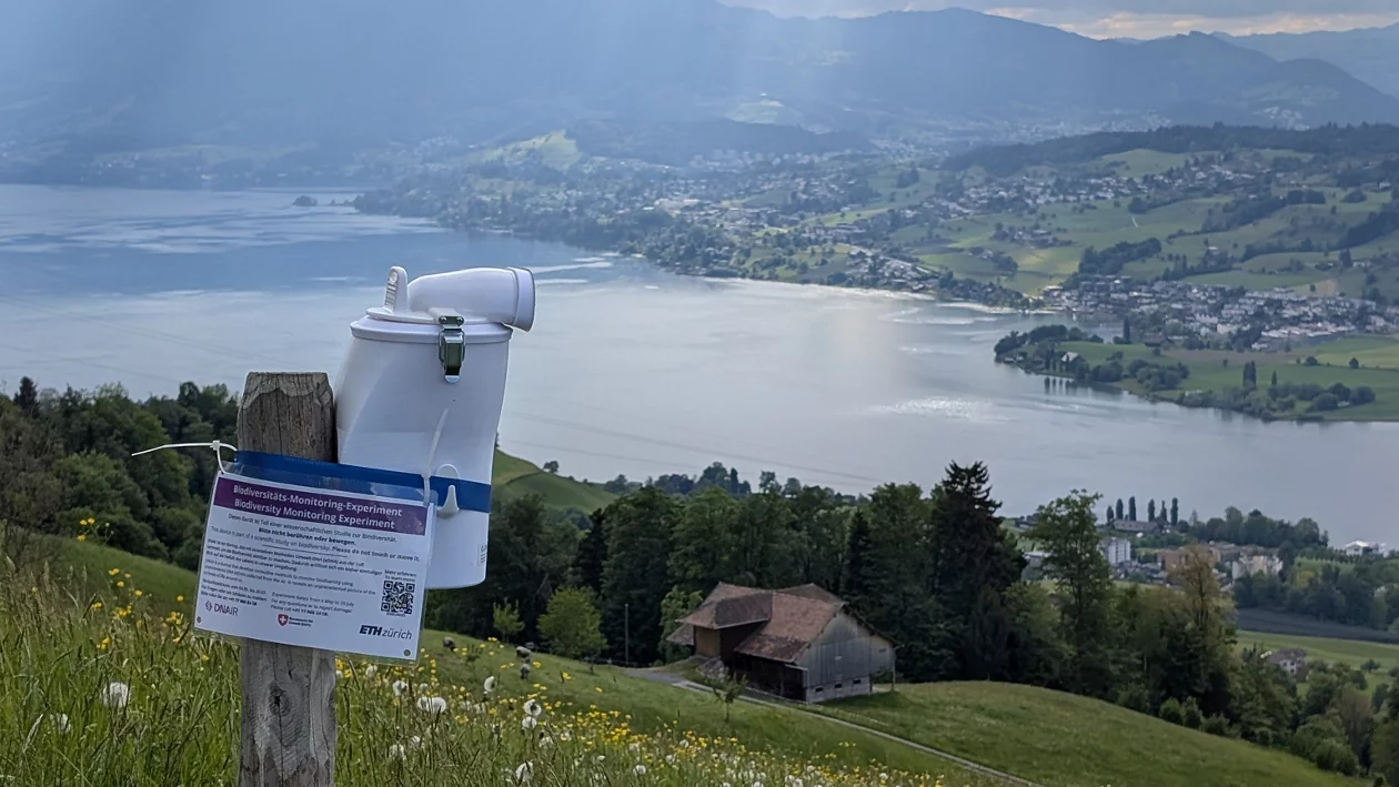 Auf einer Wiese berhalb eines Sees ist eine Messstation an einem Holzpfosten montiert.