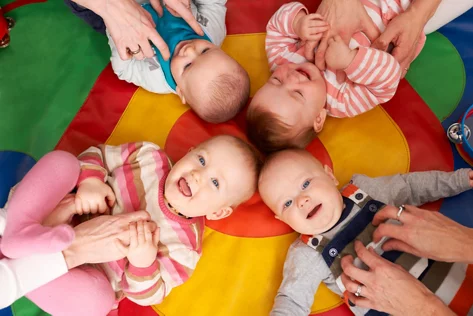 Babys liegen auf einem bunten Tuch und werden von ihren Eltern gekitzelt.