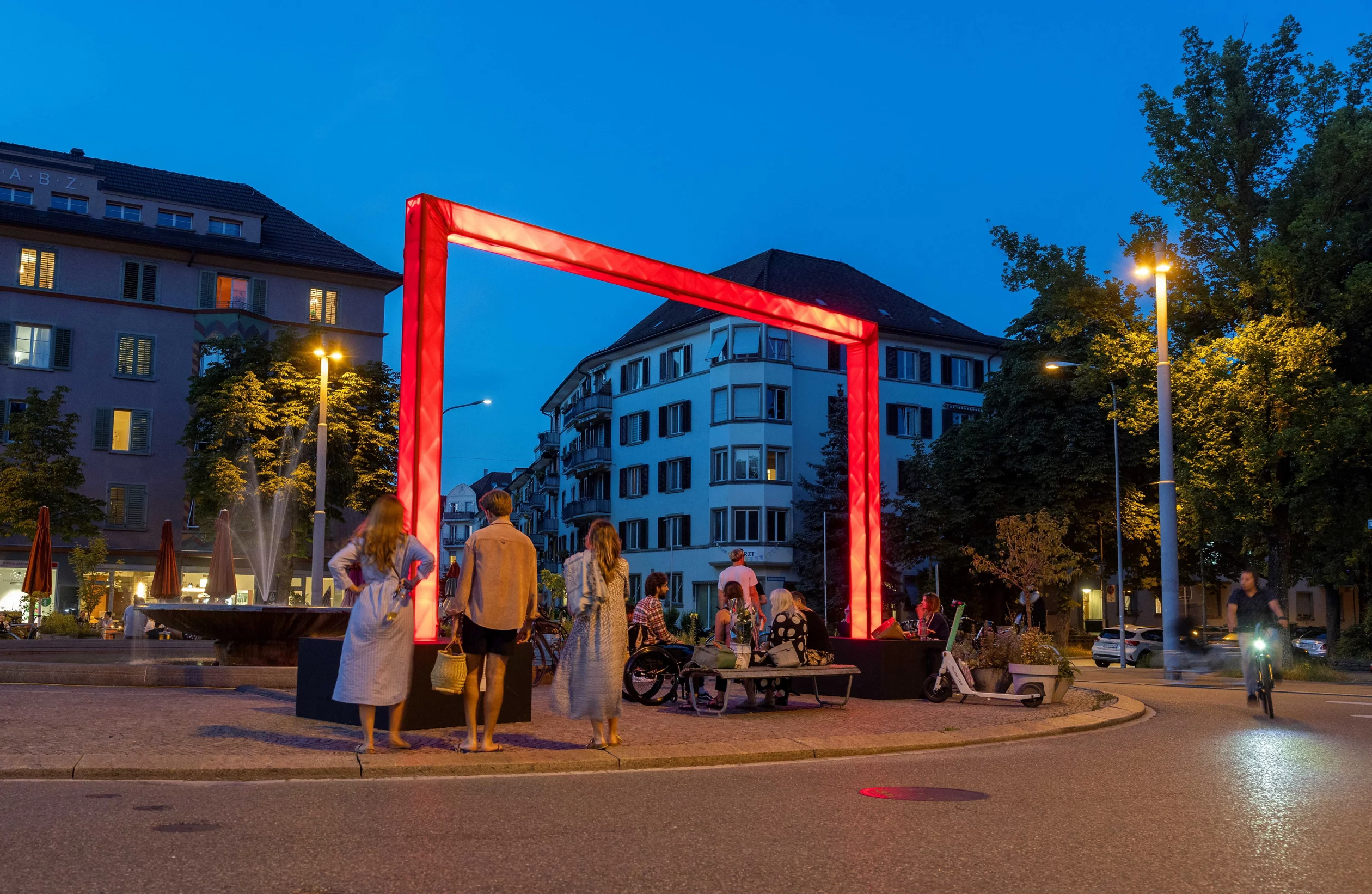 Auf dem Bullingerplatz dunkelt es ein. Vor dem Blau des Himmels leuchtet ein übergrosses rotes Tor, darum herum stehen und sitzen junge Menschen.