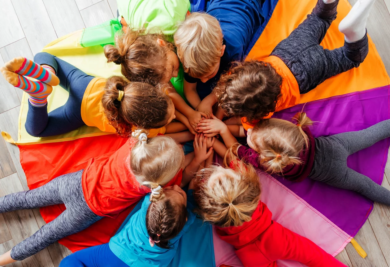 Eine Gruppe von Kleinkindern liegt auf einem bunten Regenbogentuch und legt die Hände in der Mitte übereinander.