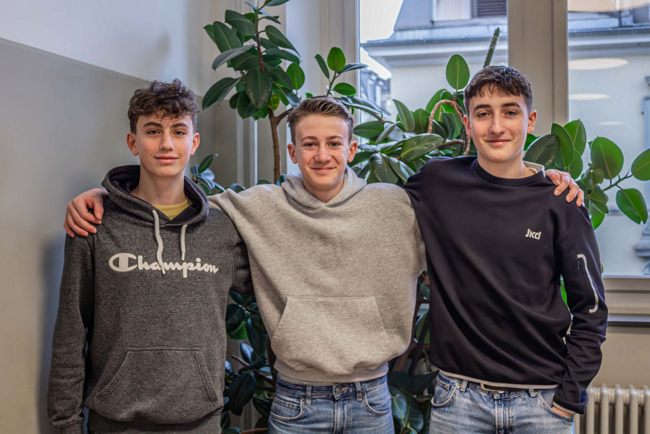 Portraitfoto von Yaniv, Matteo und Cesare vor einer Pflanze; sie haben die Arme umeinander gelegt und lachen.