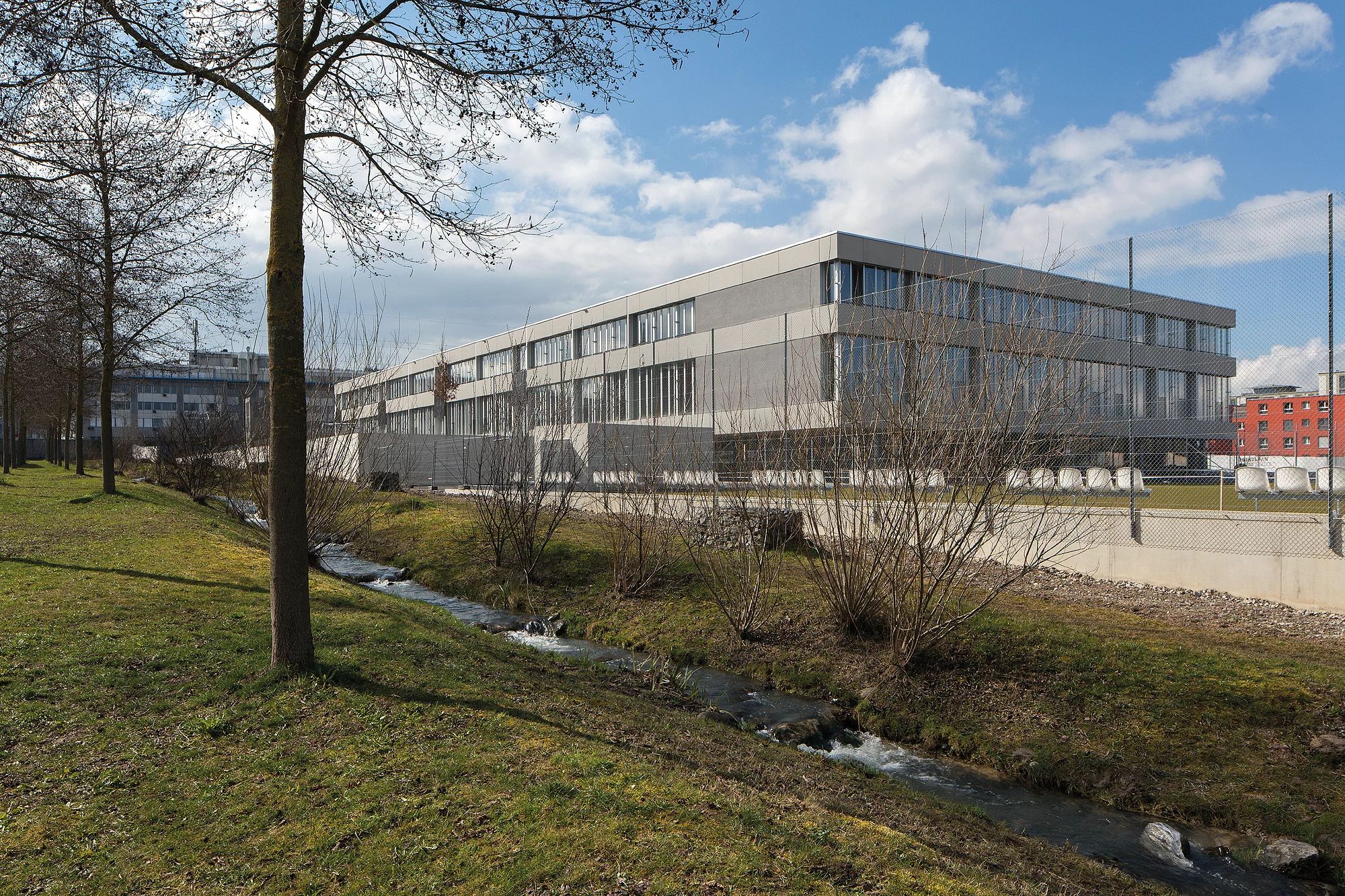 Das Foto wirft einen Blick über eine grüne Umgebung mit Bach auf die Nordfassade der Schulanlage.