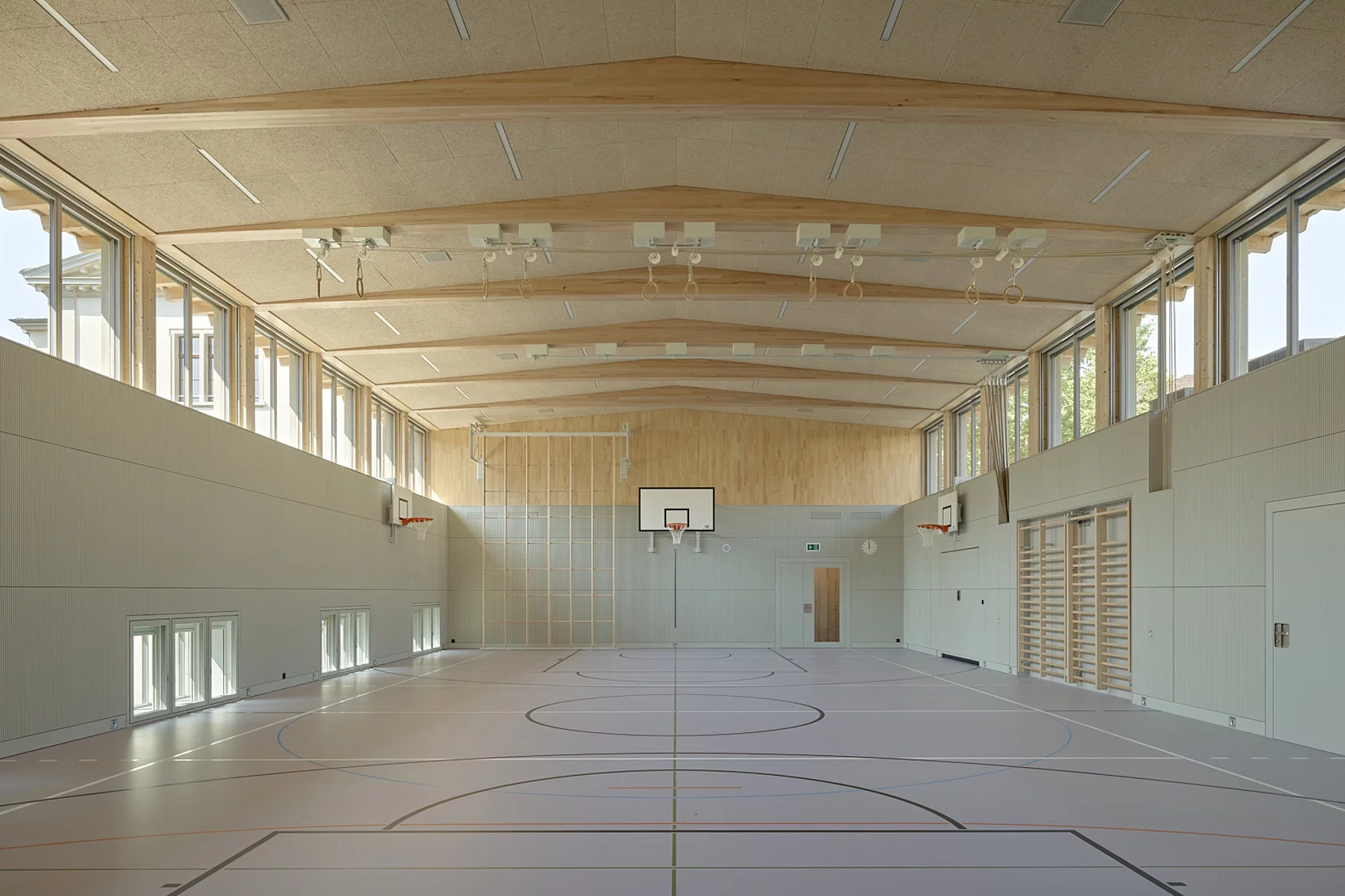 Helle, moderne Sporthalle mit Basketballkörben, Holzdach und grossen Fenstern entlang der Seitenwände.