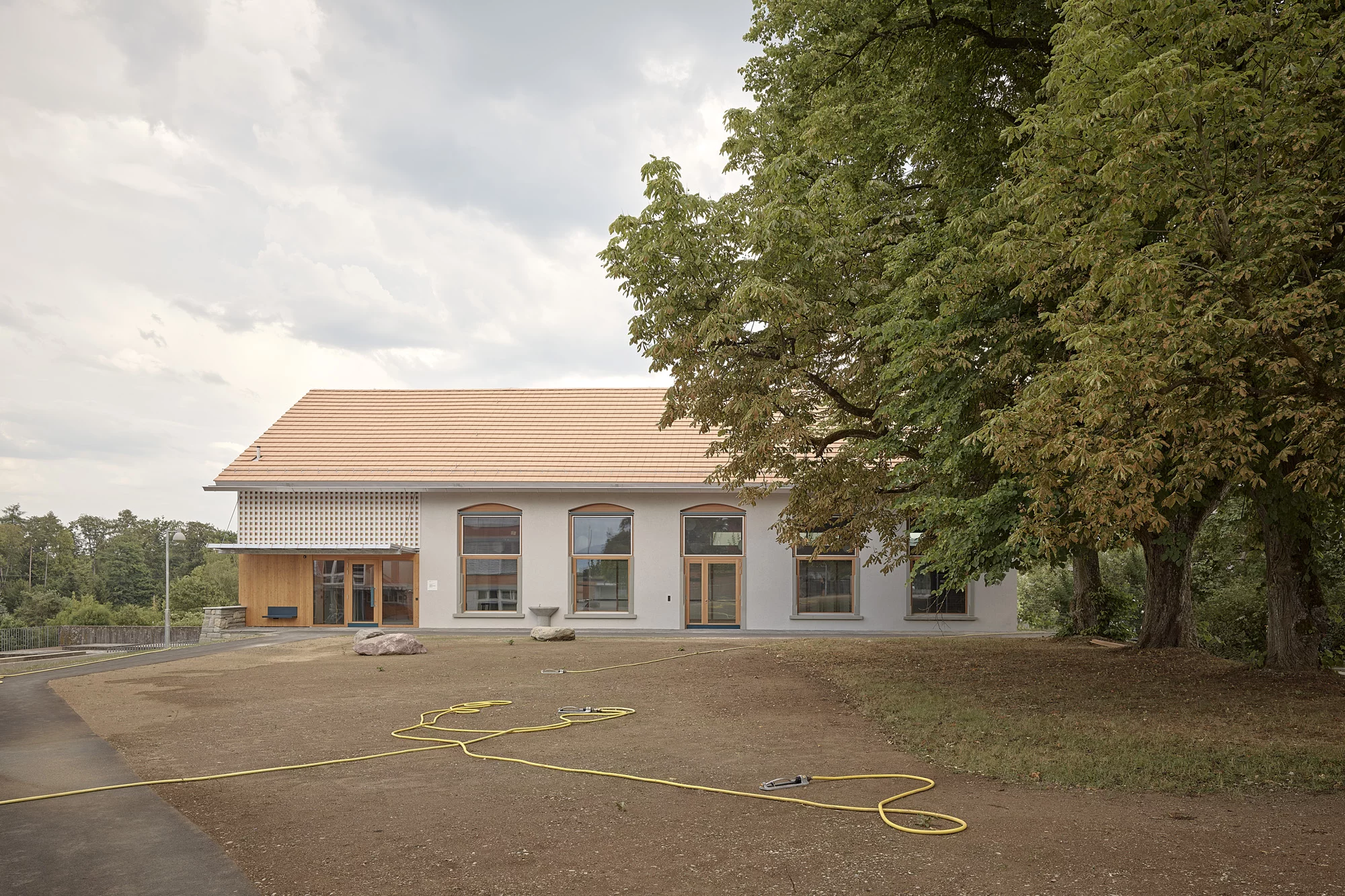 Anbau und Alte Turnhalle nach dem Umbau: Modernisiertes Gebäude mit hellem Putz und Satteldach vor grossen Bäumen.
