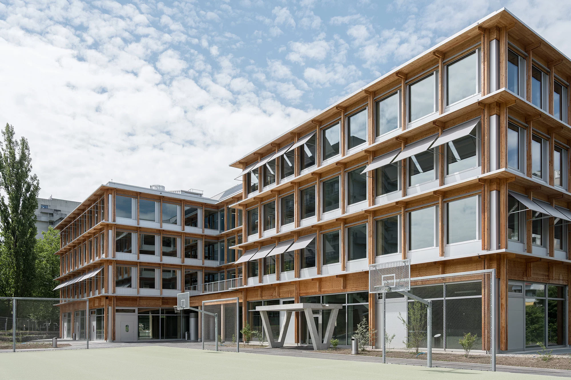 Neubau der Schulanlage Wolfsblick mit Holzfassade, grossen Fenstern und Sportplatz mit Basketballkörben im Vordergrund.