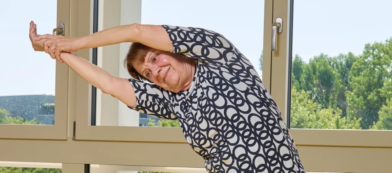 Eine ältere Frau dehnt sich vor einem Fenster. 