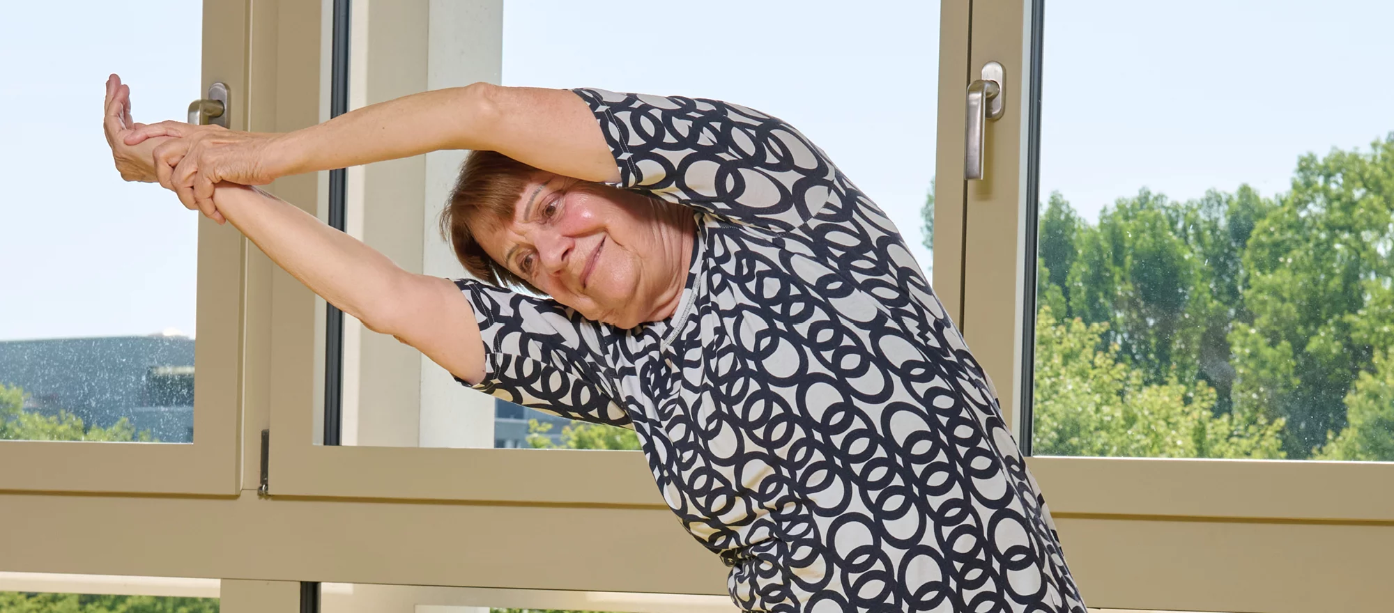 Eine ältere Frau dehnt sich vor einem Fenster. 