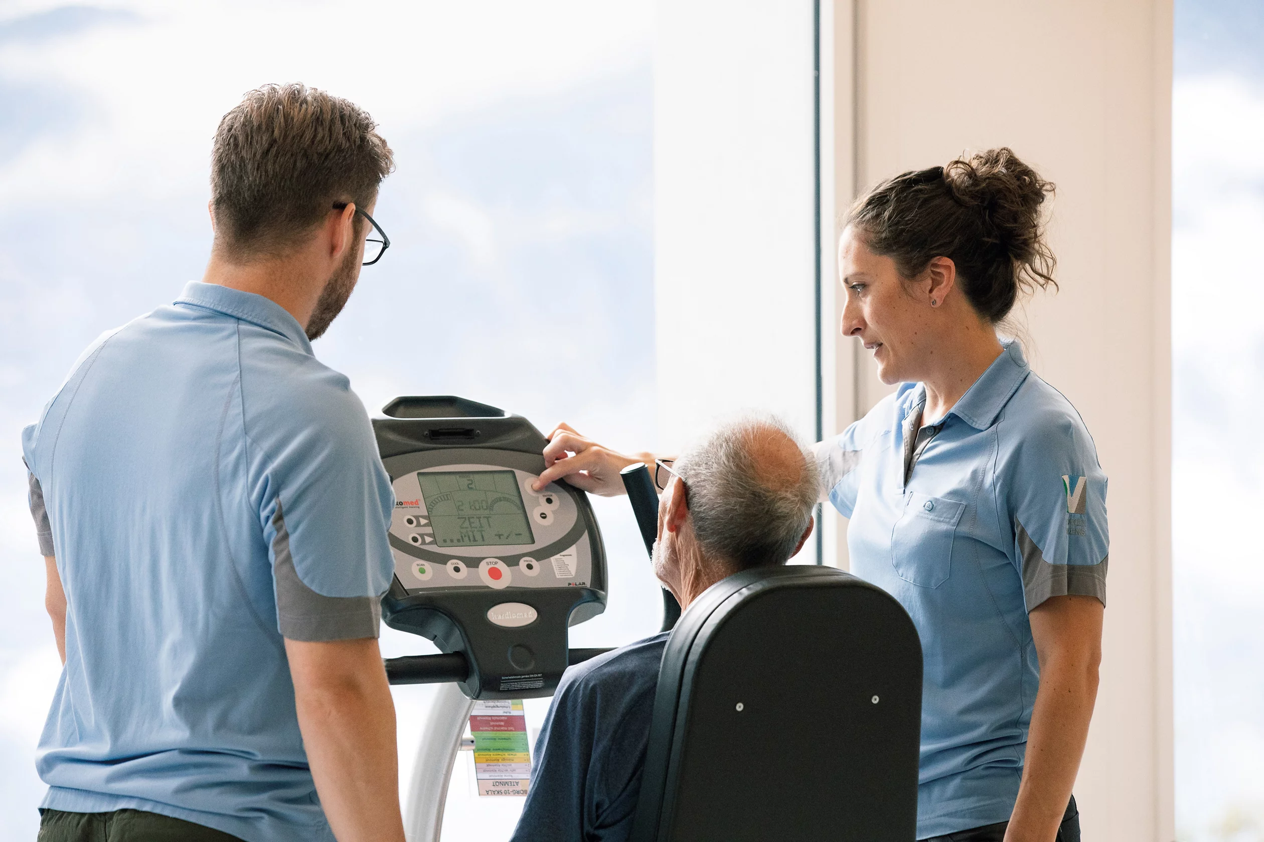 Älterer Mann auf dem Fahrrad der Physiotherapie und zwei Therapeutinnen daneben