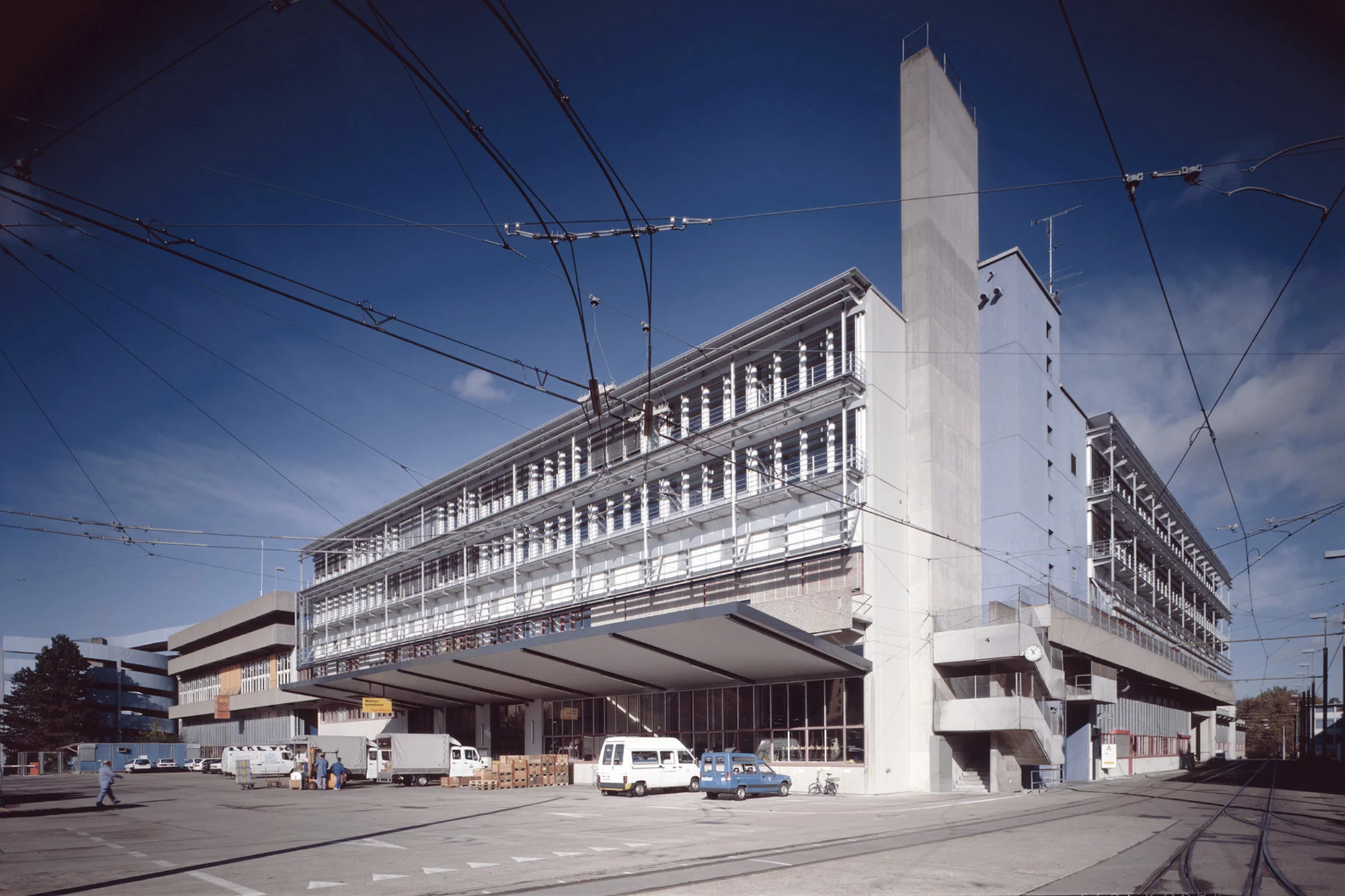 Grosses helles Gebäude der VBZ Zentralwerkstatt an einer Strasse mit Tramleitungen im Vordergrund.