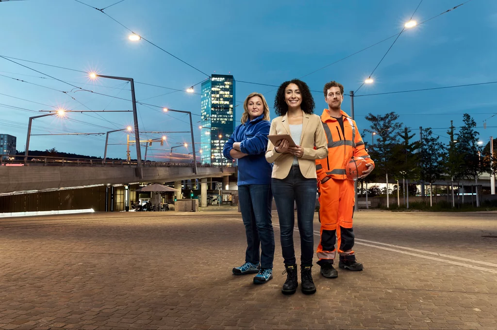 Drei Mitarbeitenden vom erz in Arbeitskleidung in Dämmerung vor Prime Tower stehend.