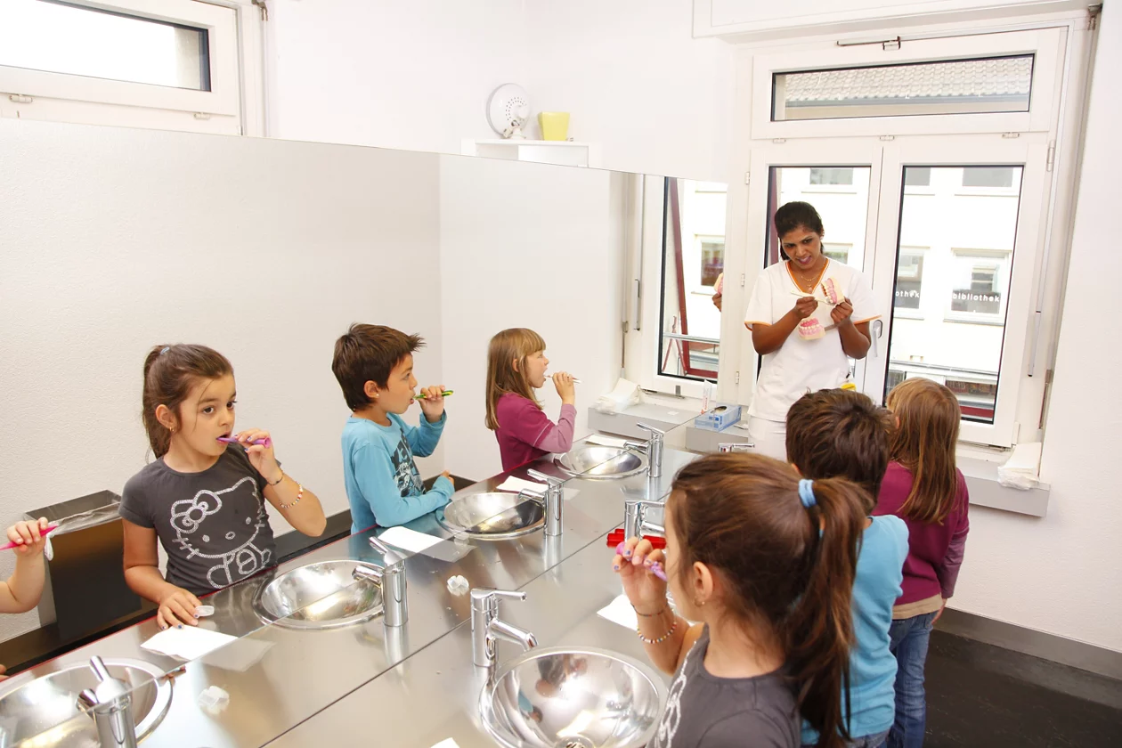 Zahnputzinstruktorin zeigt vor wie die Kinder die Zähne putzen sollen. Kinder stehen am Lavabo und putzen sich die Zähne.