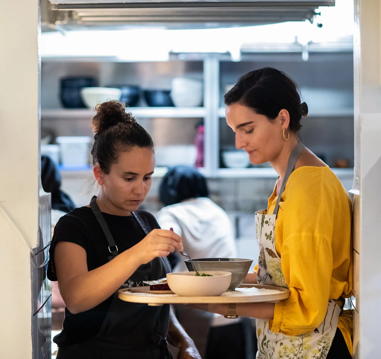 Zwei Frauen stehen in der Tür zur Restaurantküche, eine Mitarbeiterin hält ein Tablett mit Speisen, Zizi Hattab prüft sie.
