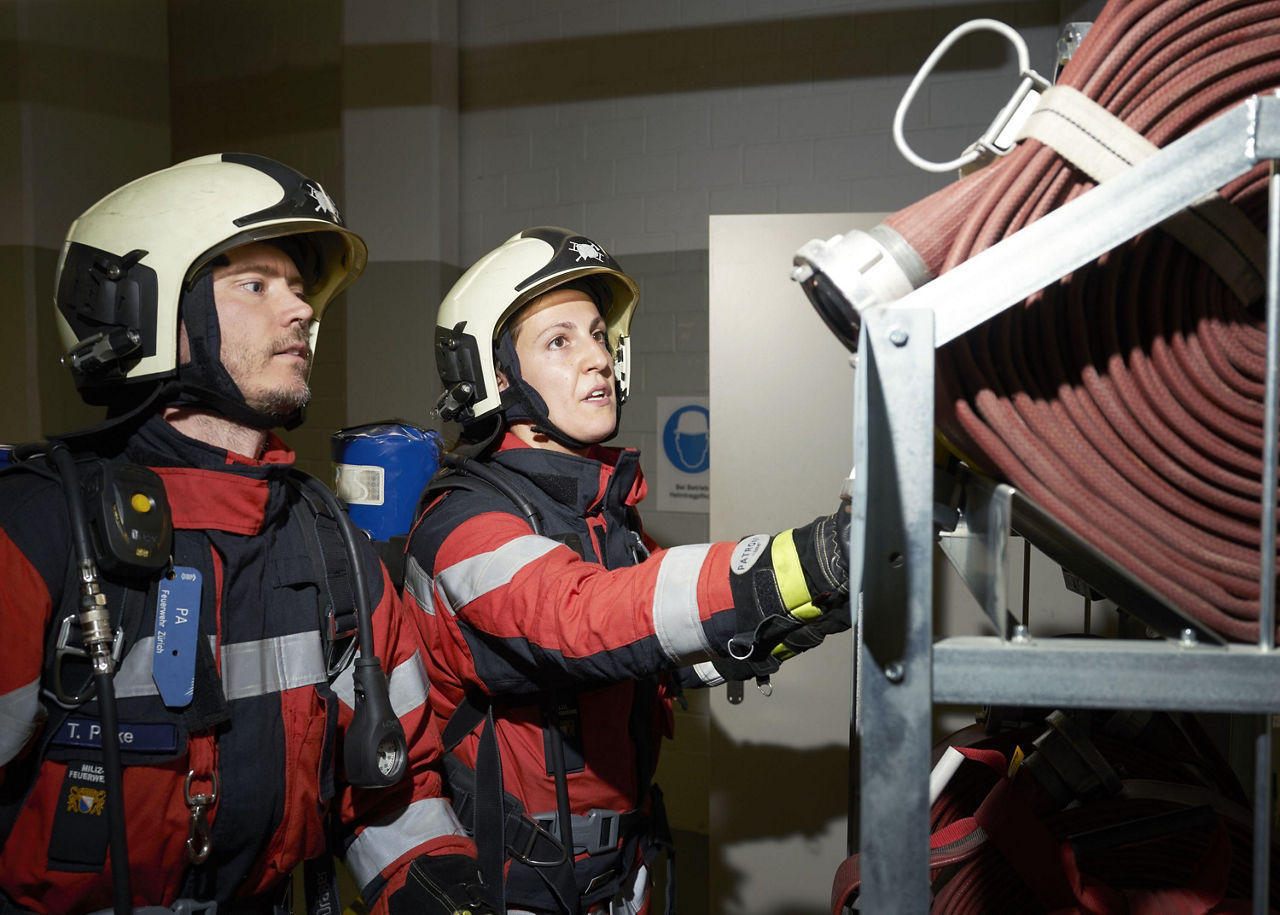 Zwei Milizfeuerwehrleute rollen einen Löschschlauch ab. 