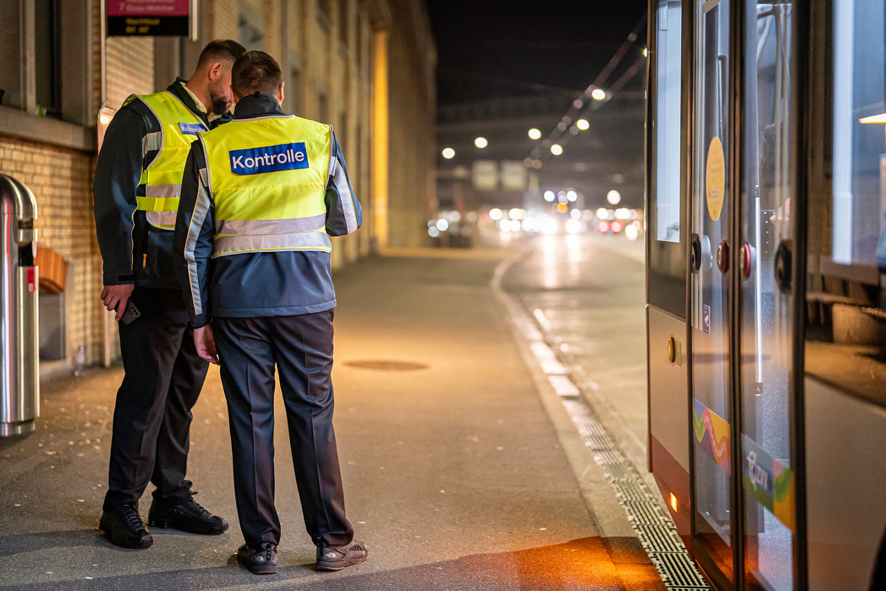 Zwei Kontrolleure in gelben Warnwesten mit der Aufschrift ‚Kontrolle‘ stehen abends an einer Tramhaltestelle neben einem haltenden Tram und sprechen miteinander.