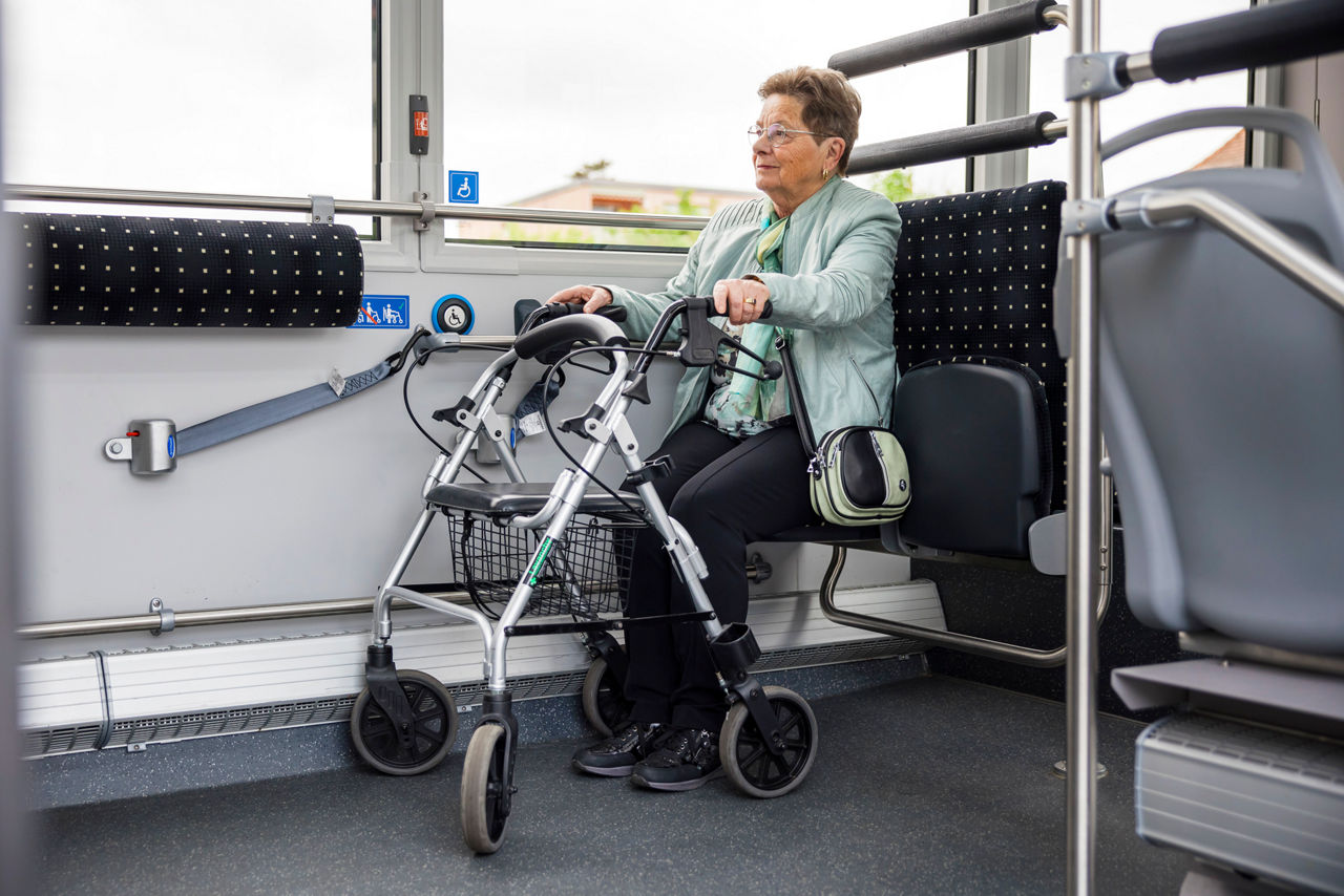Eine Seniorin sitzt auf dem Klappsitz im Bus und hält ihren Rollator fest.